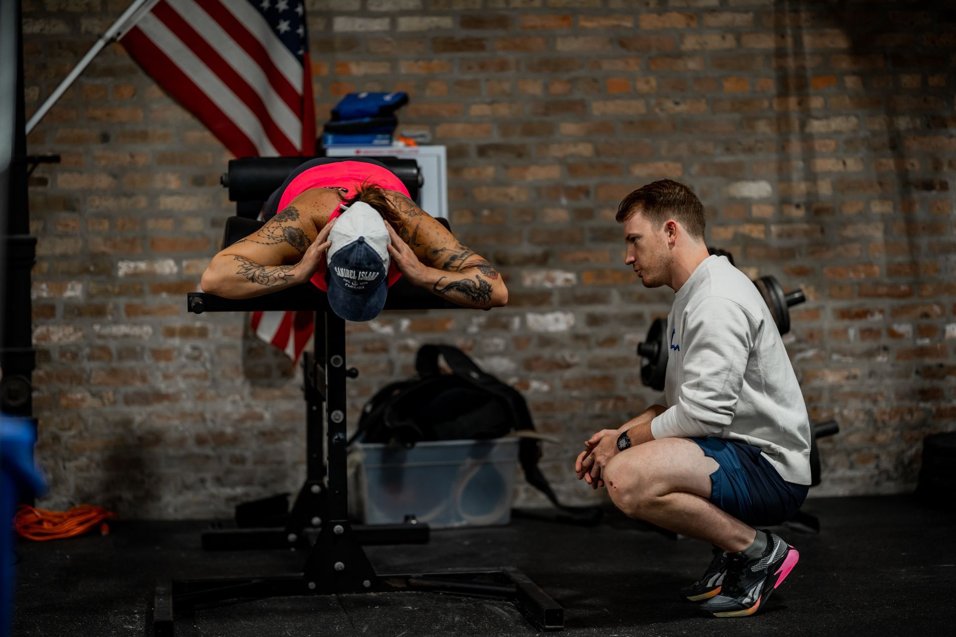A person with tattoos performs a back exercise on a hyperextension bench while being observed by a coach in a gym.