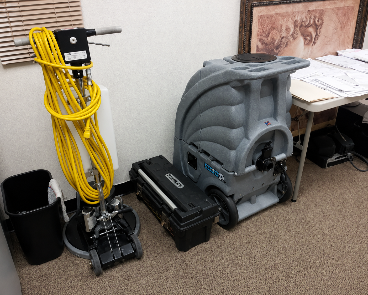 Yellow carpet cleaner and gray floor scrubber in a room with cleaning supplies and furniture.