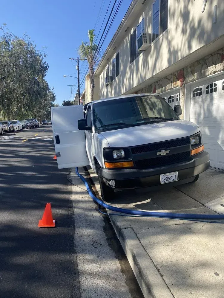 White van parked at curb with open side door, orange cone, and hoses on a sunny street.