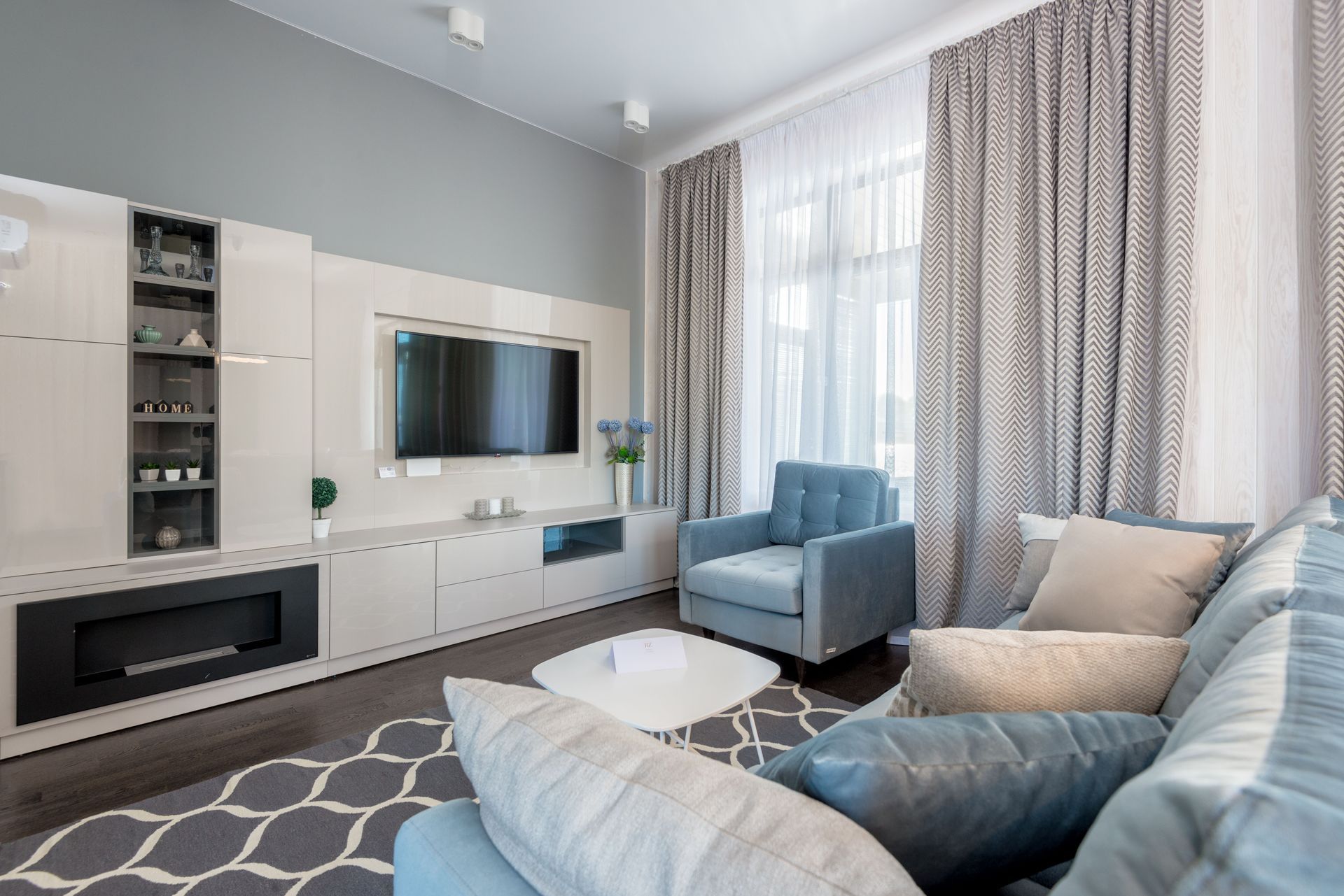 Bright modern living room with blue sofa, armchair, wall-mounted TV, and large windows with curtains