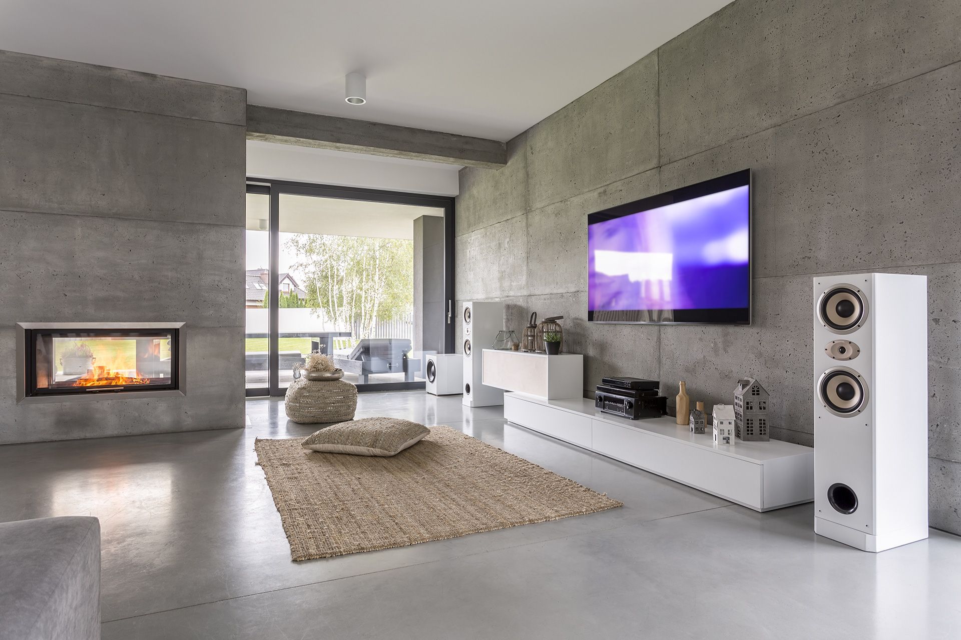 Modern living room with concrete walls, fireplace, large TV, white speakers, and sliding glass door.