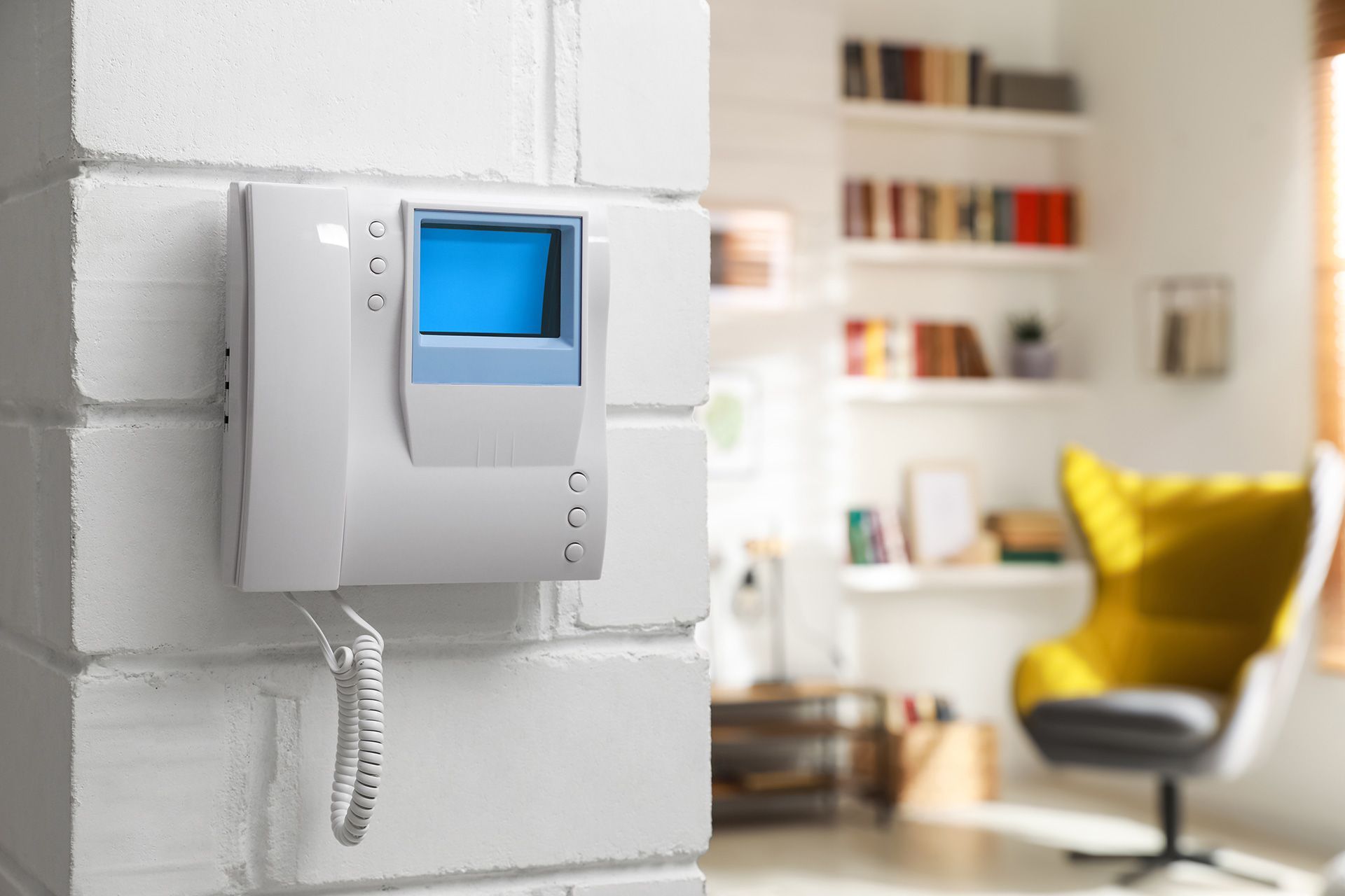 White intercom device on a brick wall, interior scene with a bookshelf and yellow chair.