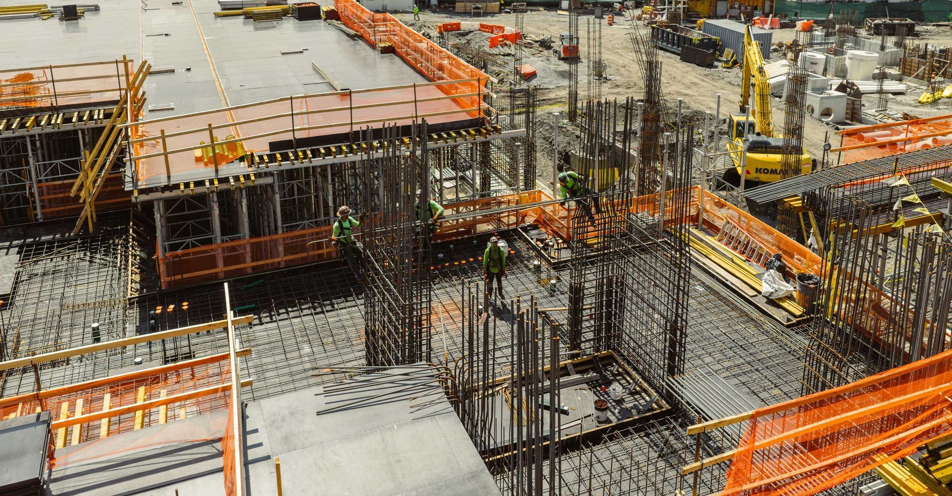 Construction site with rebar, formwork, safety fencing, and a yellow excavator.