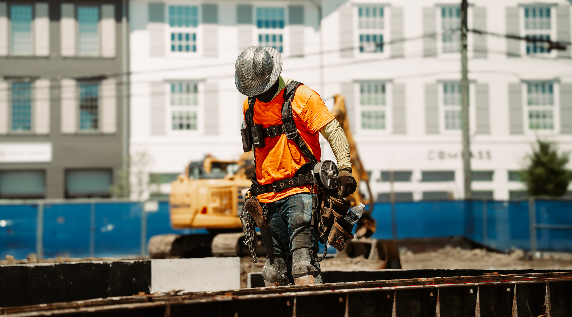 Construction worker in orange vest and helmet walks over a construction site barrier; excavator in the background.