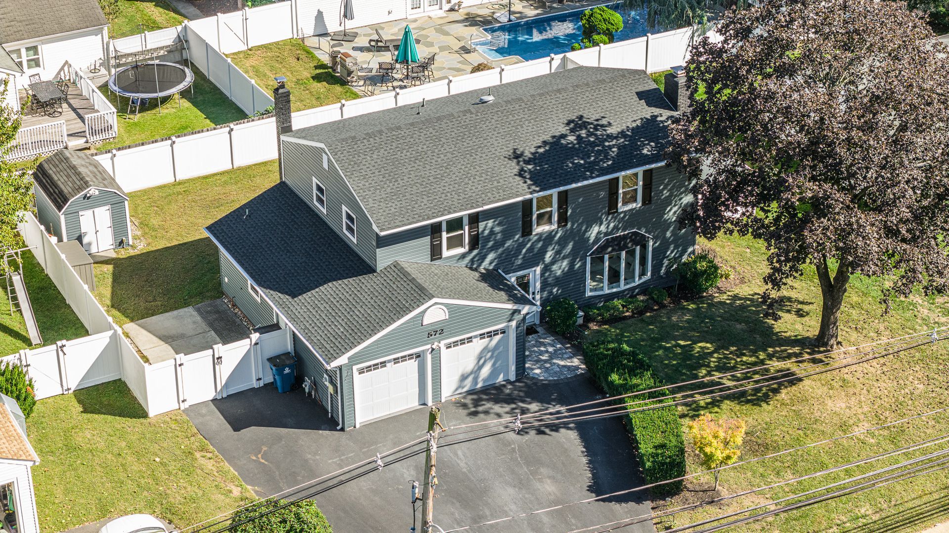 Two-story gray house with attached garage, driveway, and white fence, viewed from above on a sunny day.