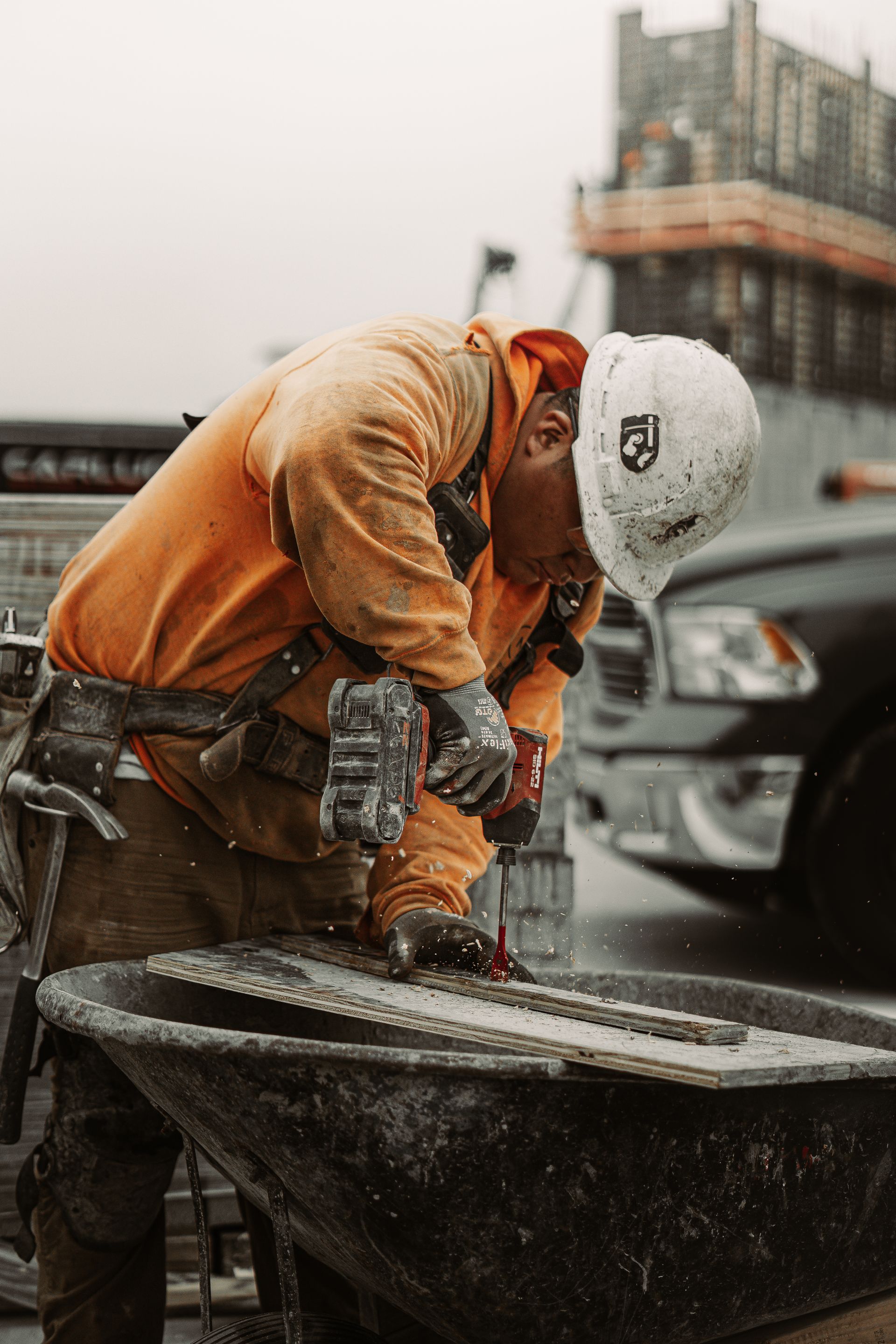 Construction worker in orange suit and hard hat using a drill near a wheelbarrow.