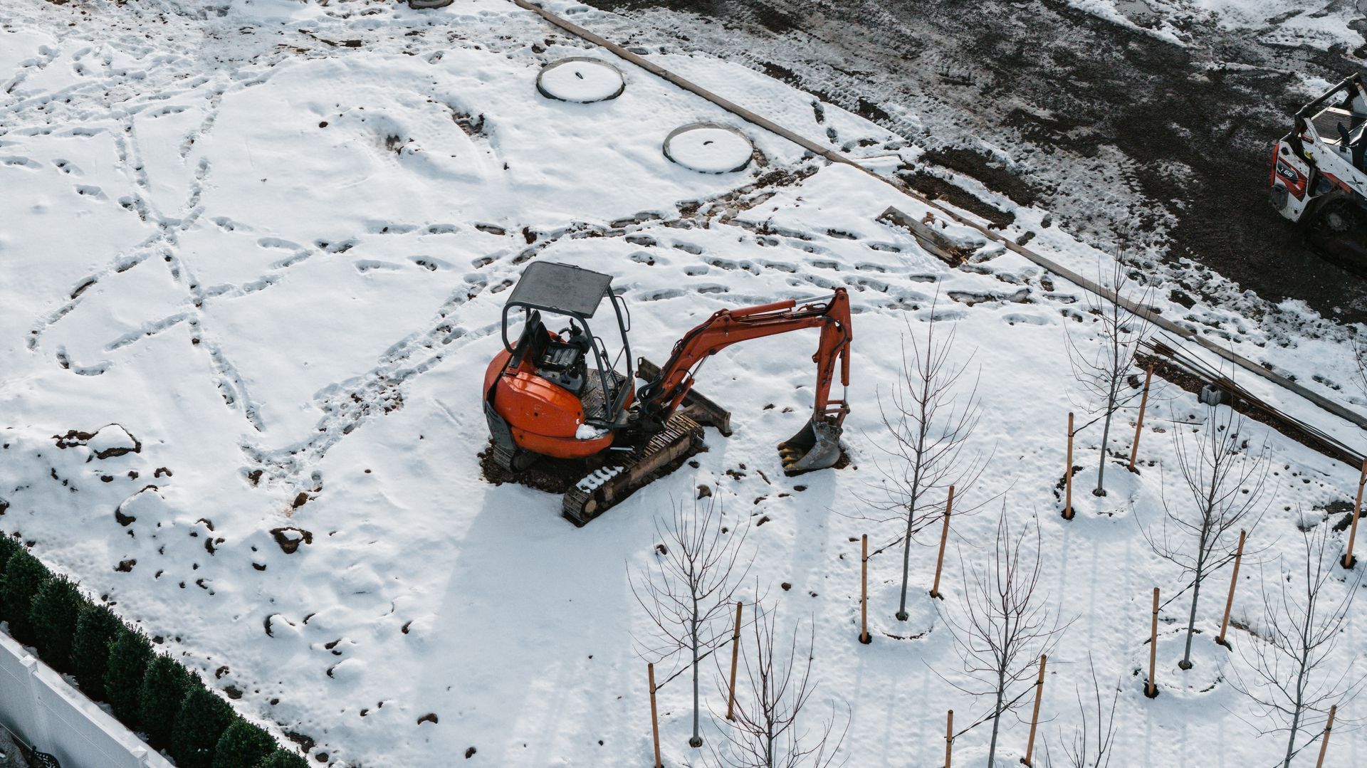Orange excavator in a snow-covered construction site, near bare trees and a concrete structure.