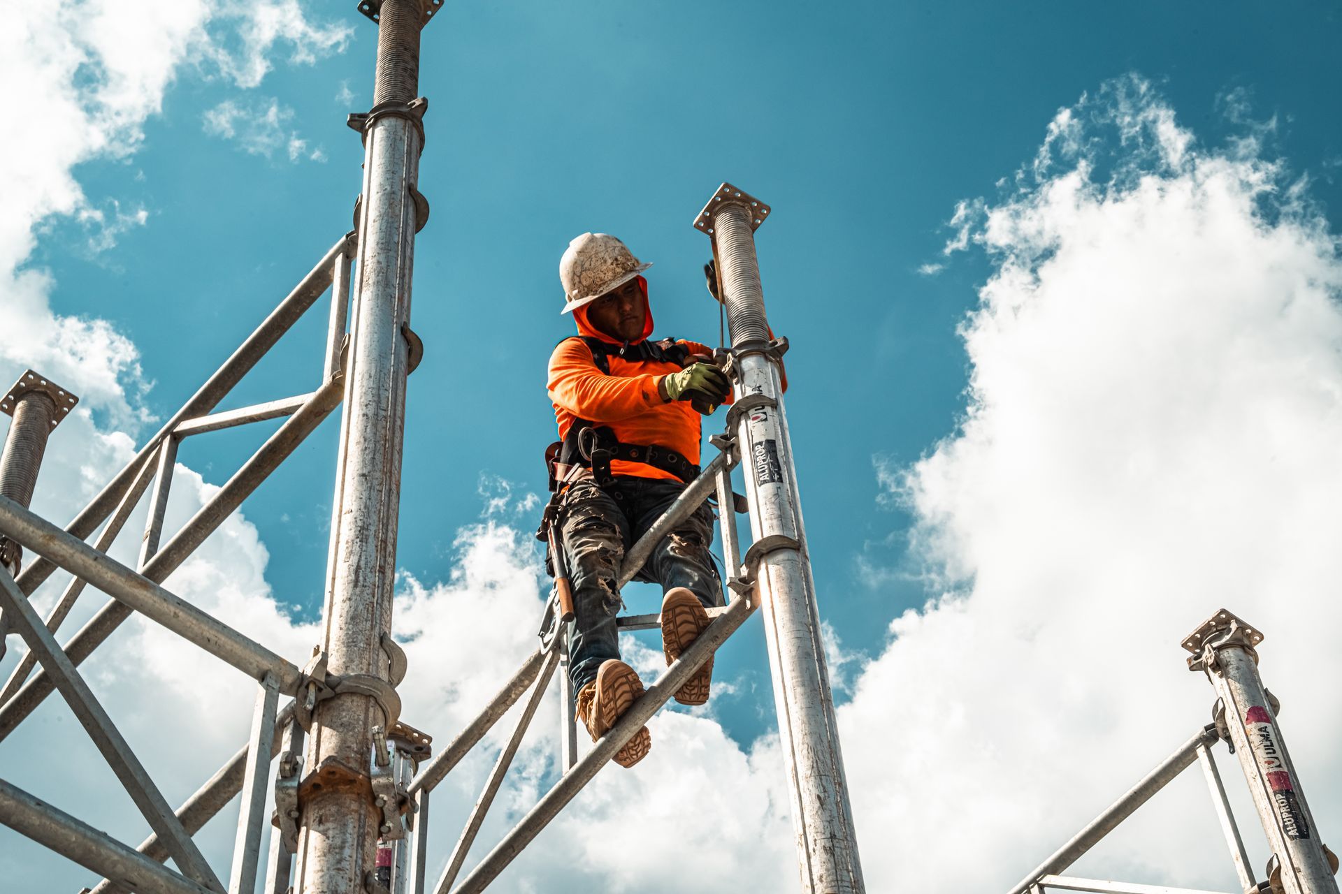 Construction worker on scaffolding, assembling metal structure against a blue sky with clouds.