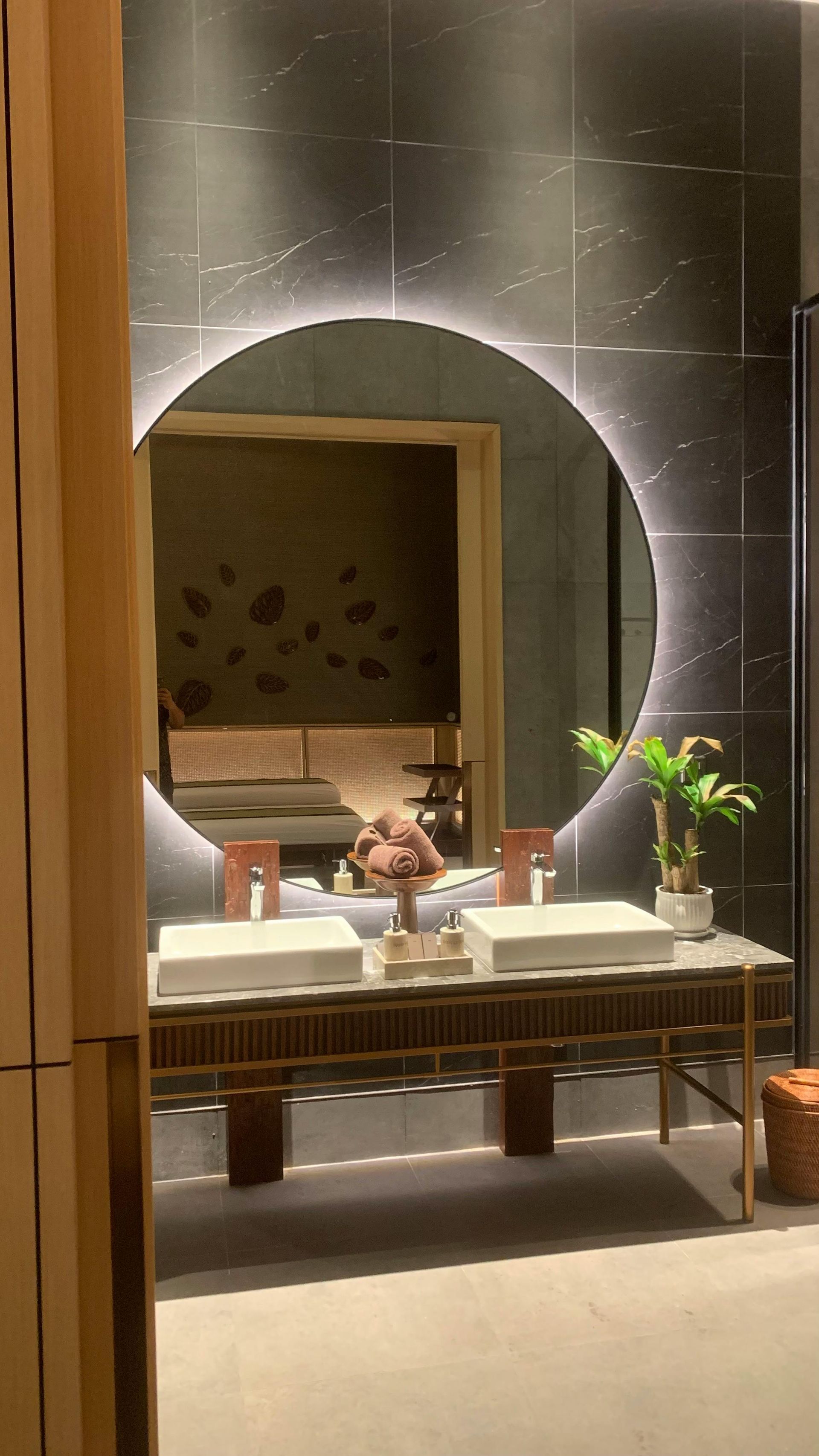 Bathroom with large round mirror, double sinks, and dark tile walls.