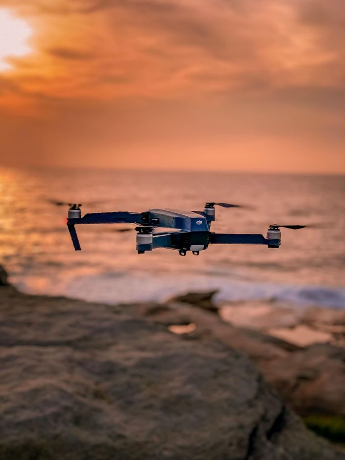 Drone in flight over a rocky coastline at sunset; orange sky, ocean waves.