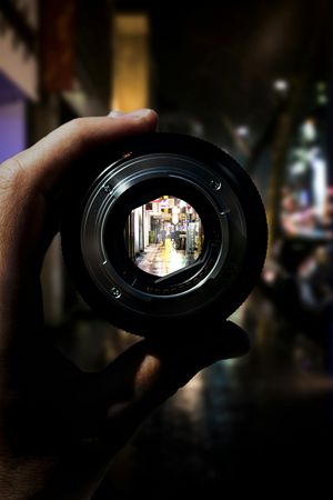 Hand holding a camera lens, with a blurry street scene visible through it.