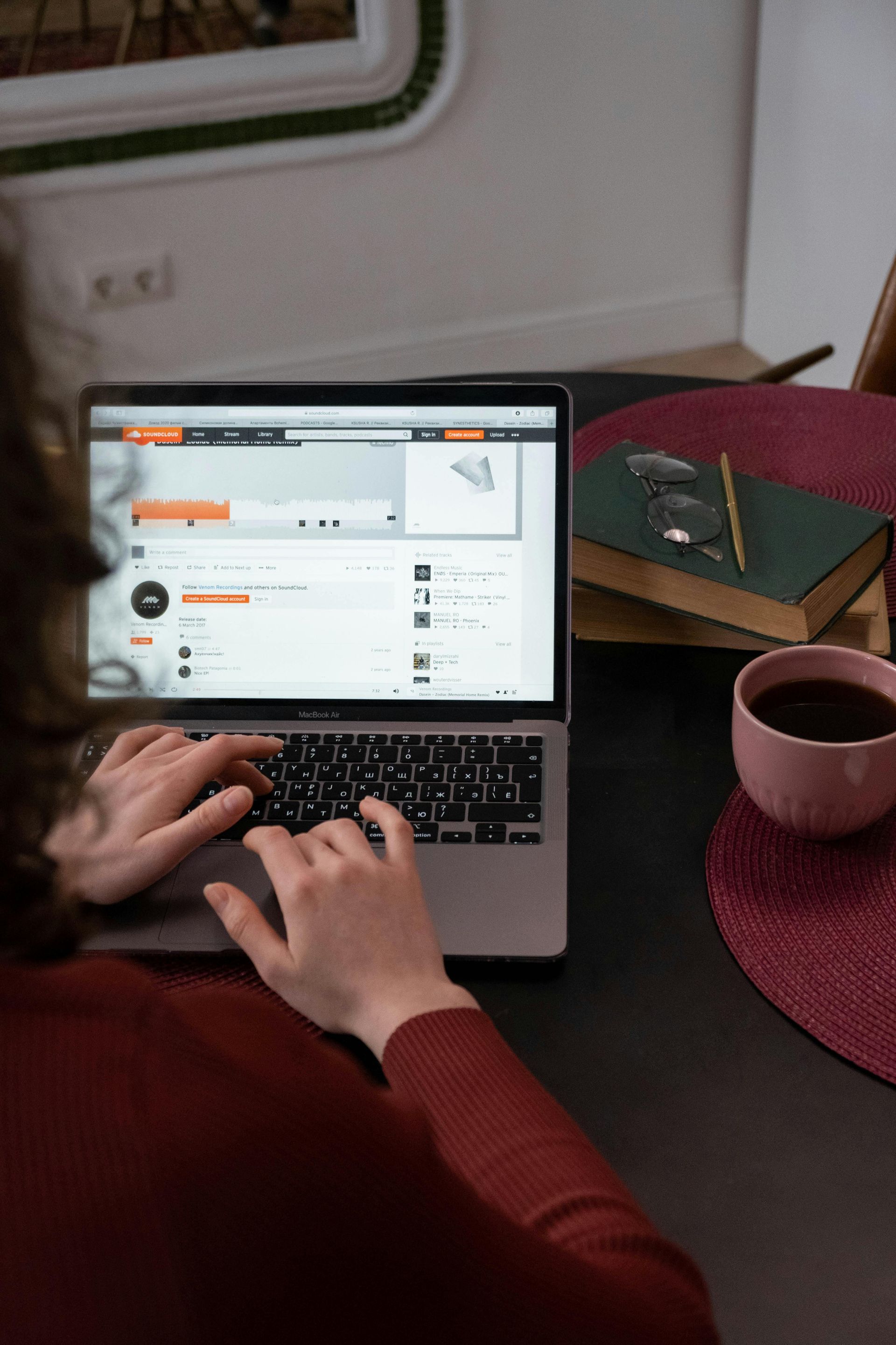Person typing on laptop, viewing website. Desk with coffee, book, and glasses. Dark clothing, warm tones.