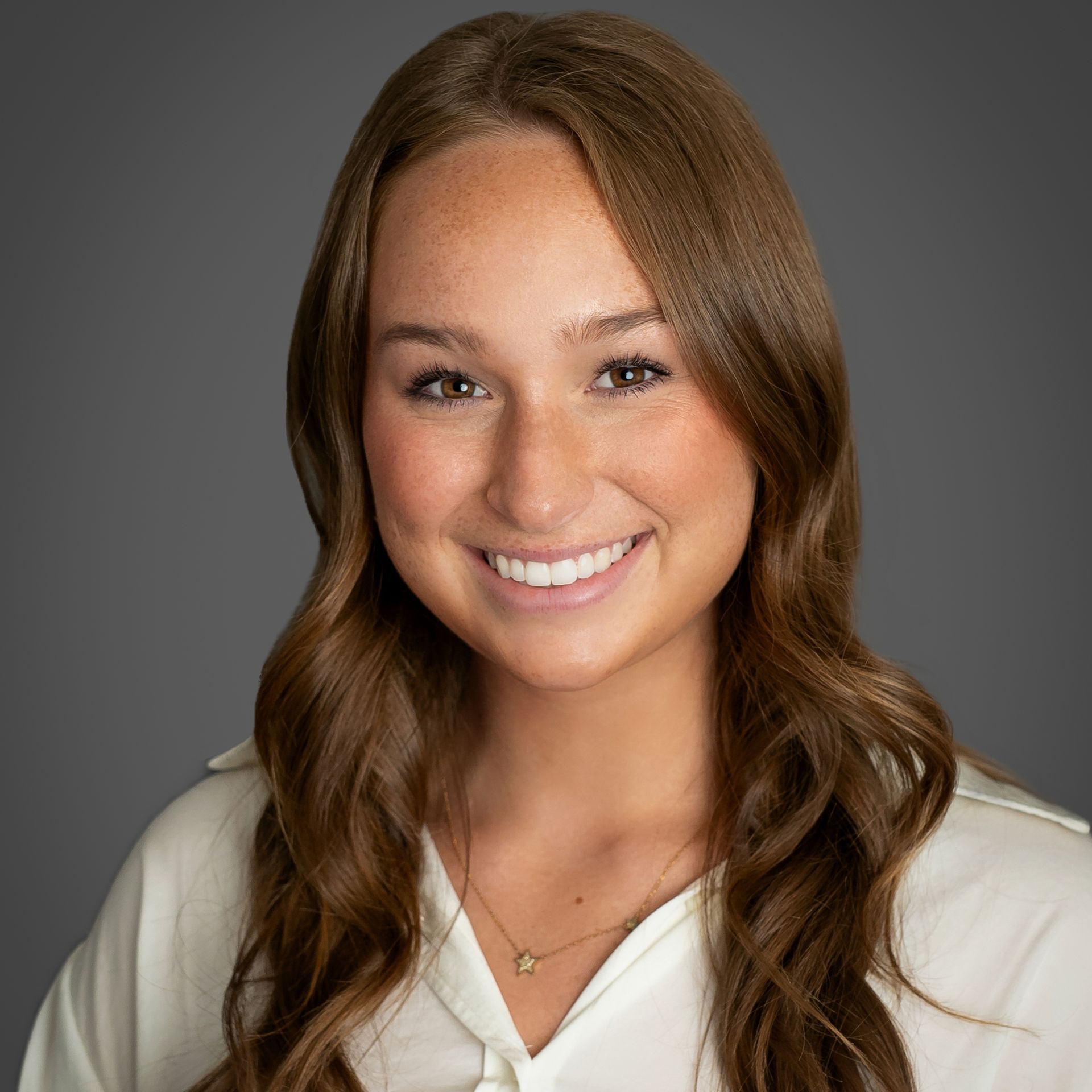 A woman with long brown hair is wearing a white shirt and smiling for the camera.
