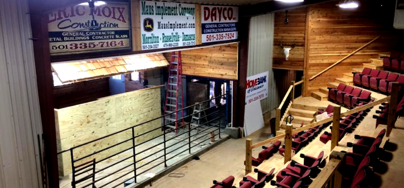 An indoor auction arena featuring tiered red seating, a plywood staging area, metal fencing, and overhead sponsor signs.