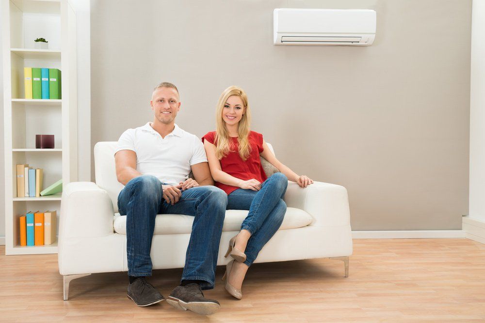 Young Happy Couple Sitting On White Sofa — Bear Air Conditioning In Saratoga, NSW