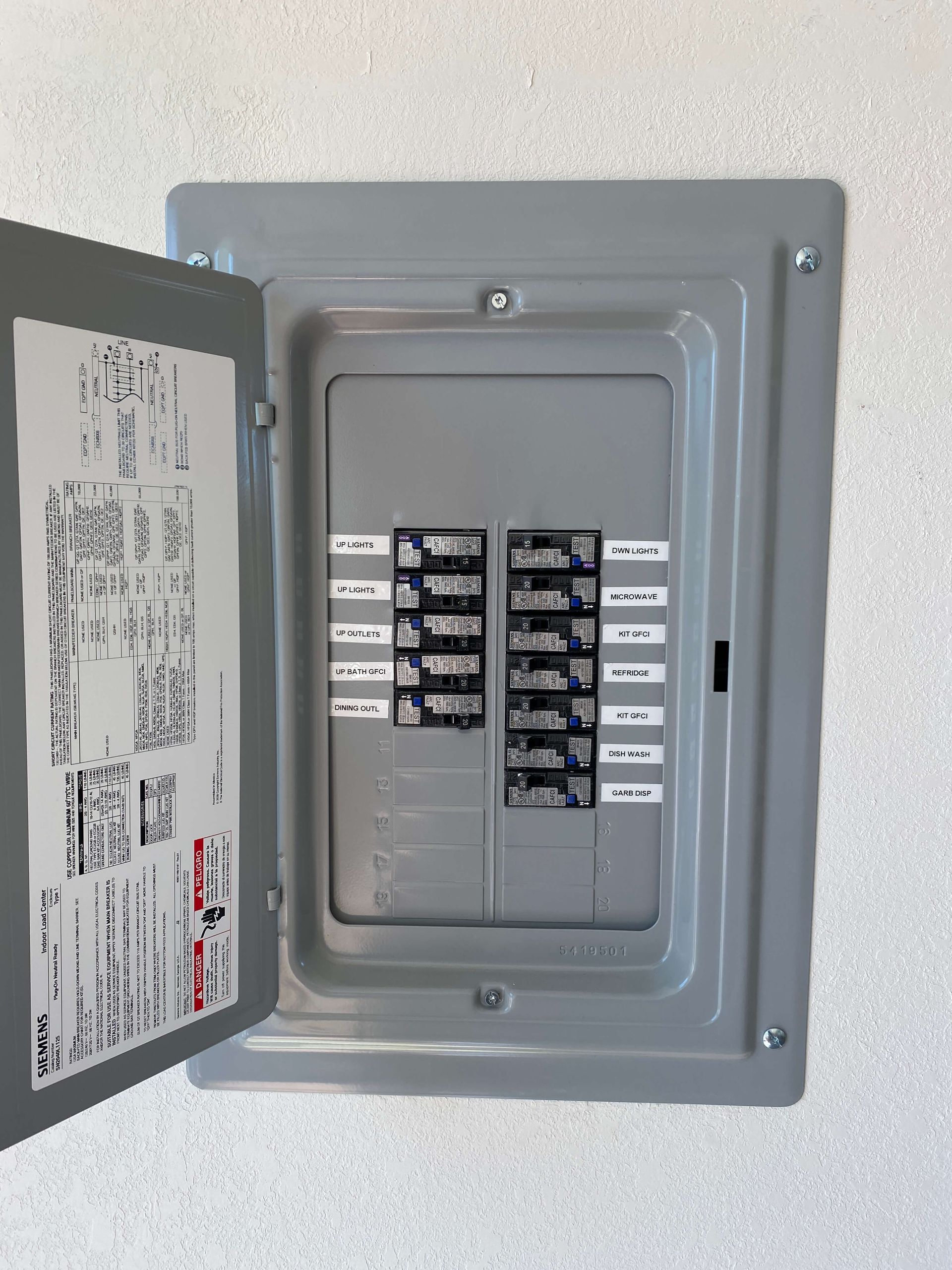 Open gray electrical breaker box with circuit breakers and labels on a white wall.
