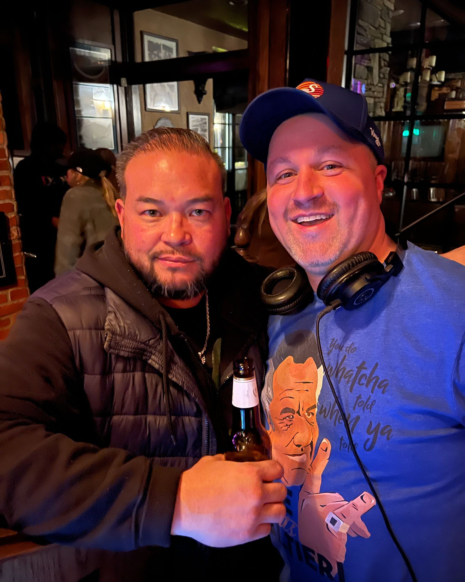 Two men smiling, one holding a beer, inside a bar. One wears a blue hat and headphones.