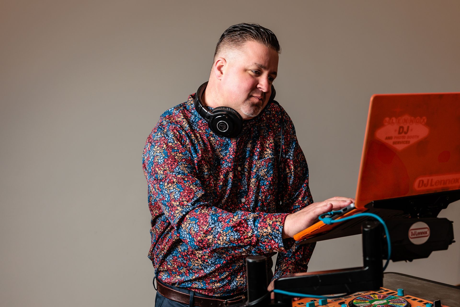 DJ working on laptop at a console, wearing headphones. Colorful shirt, orange laptop.