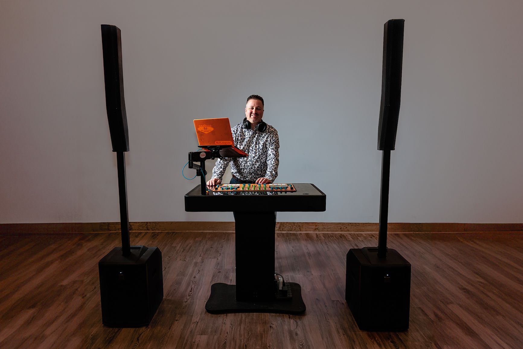 DJ at a mixing table with two tall speakers. They wear a sequined shirt and smile.