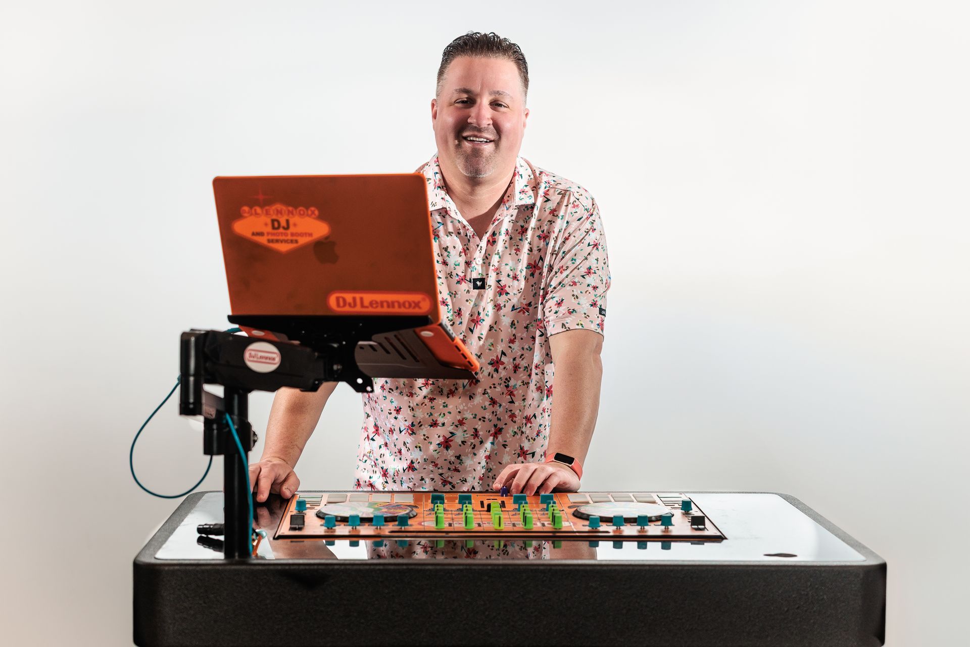 Man standing behind a table with a laptop and a colorful circuit board, smiling. White wall background.