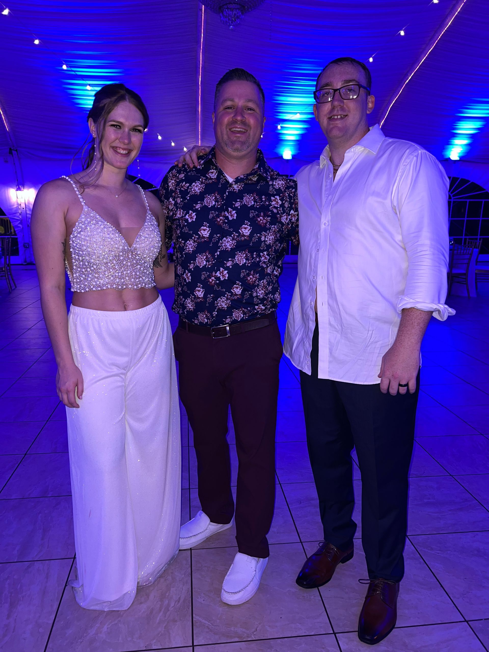 Three people pose together at an event. A woman in a sparkly top and skirt stands next to two men. Blue lighting.