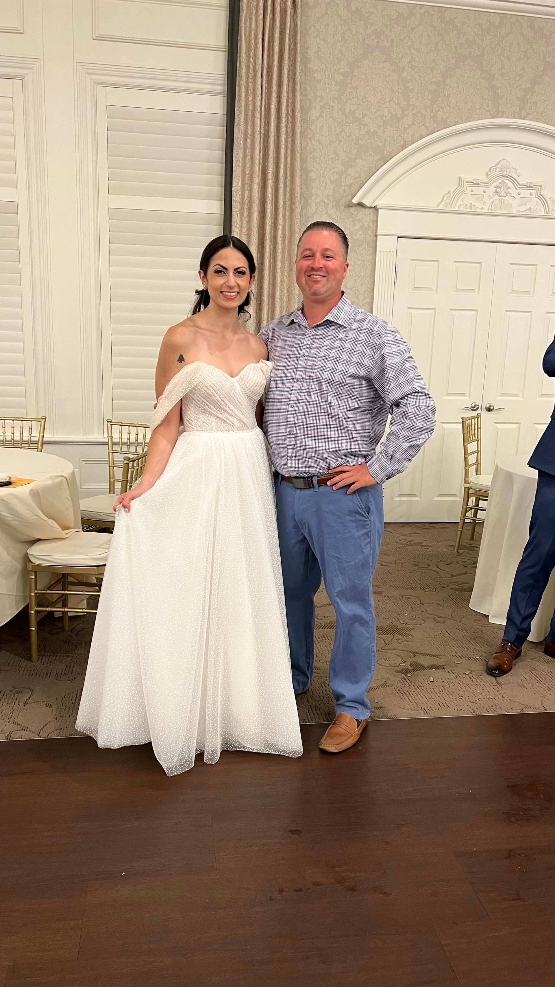 Bride in a white gown and man in a patterned shirt pose together indoors. They smile for the camera.