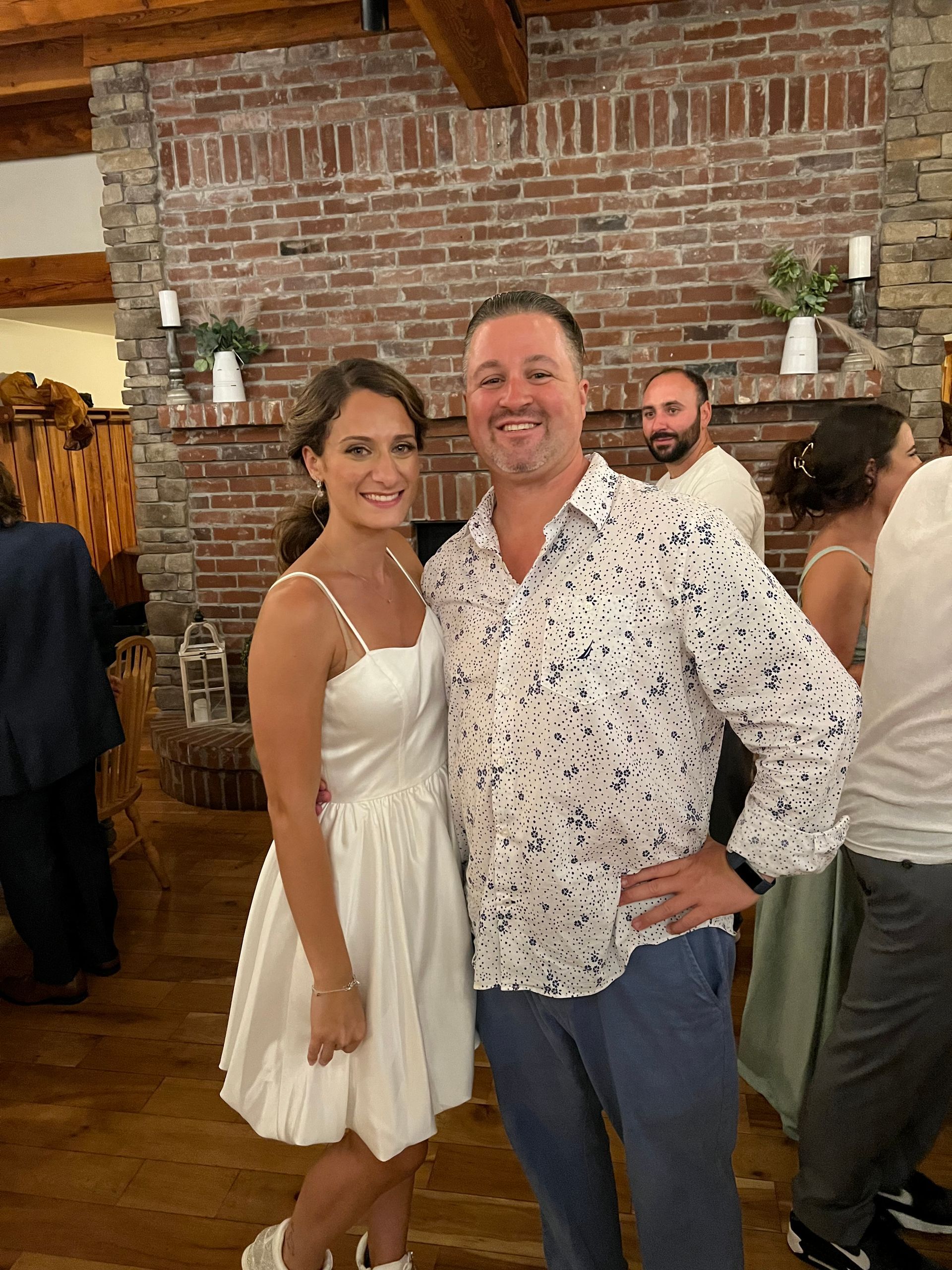 Woman in white dress and man in printed shirt smile, pose for photo in front of a brick fireplace.
