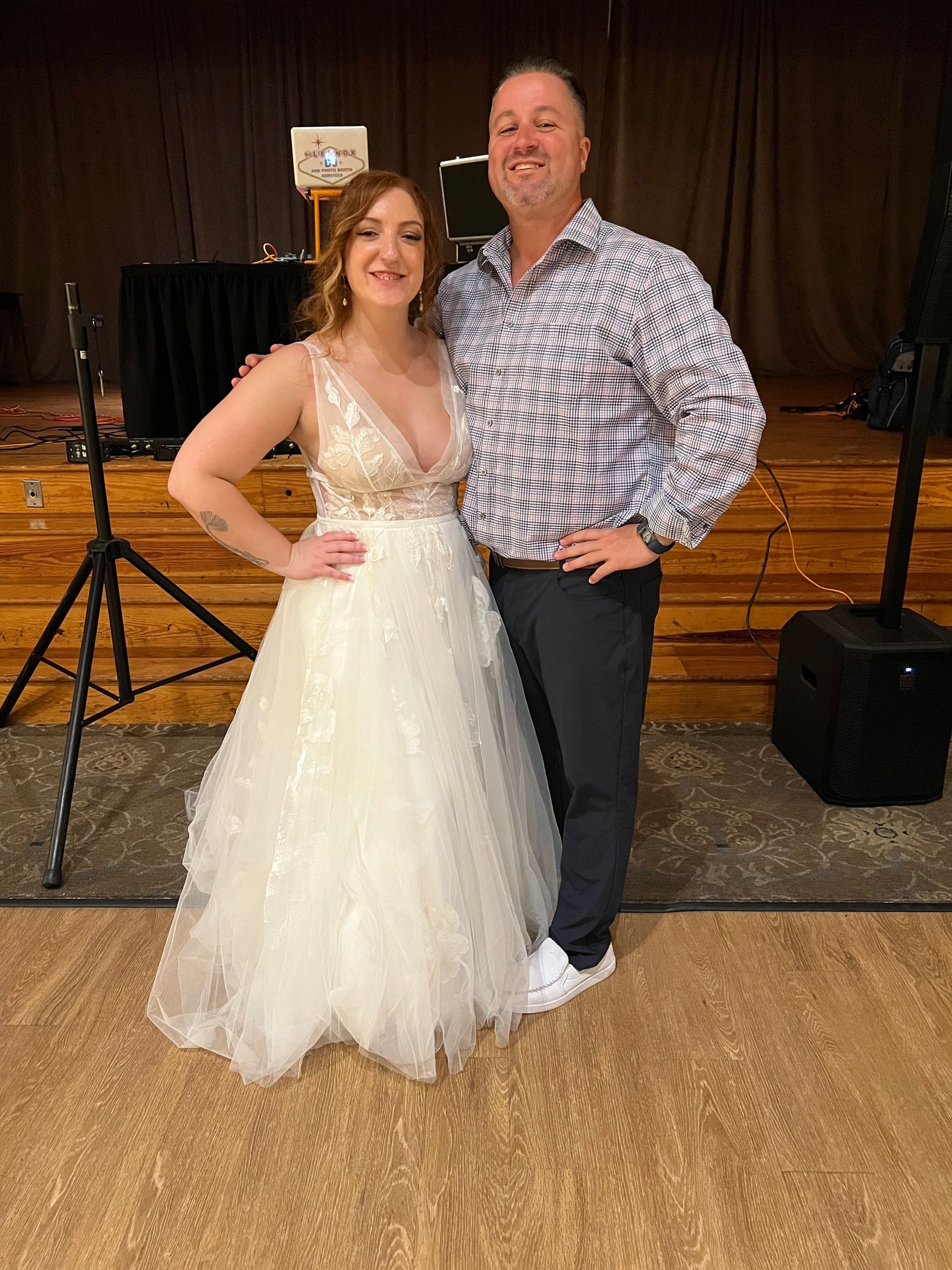 Couple posing at an event. Woman in wedding dress, man in patterned shirt. Stage background.