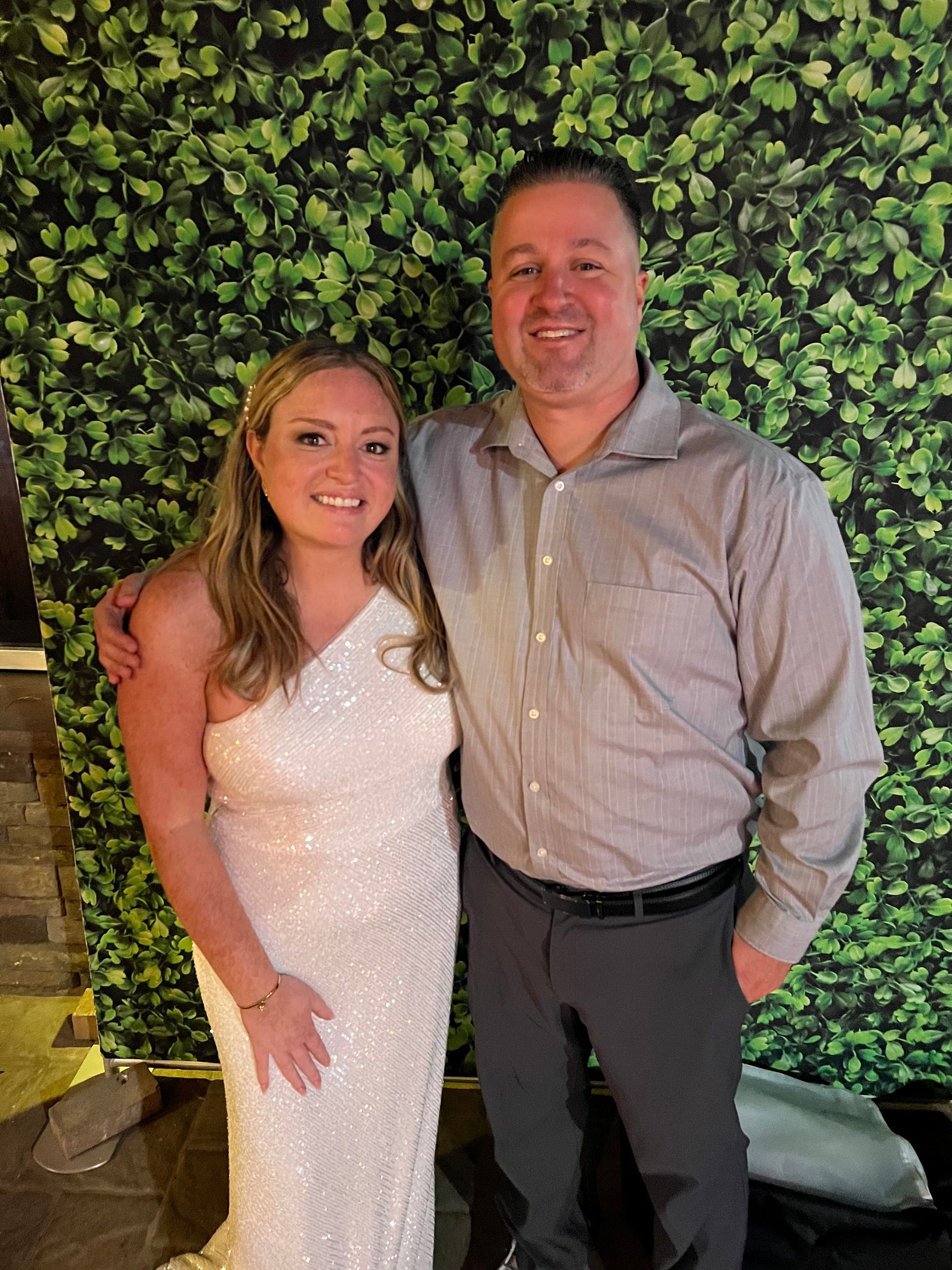 Woman in white dress and man in gray shirt pose together; green foliage backdrop.