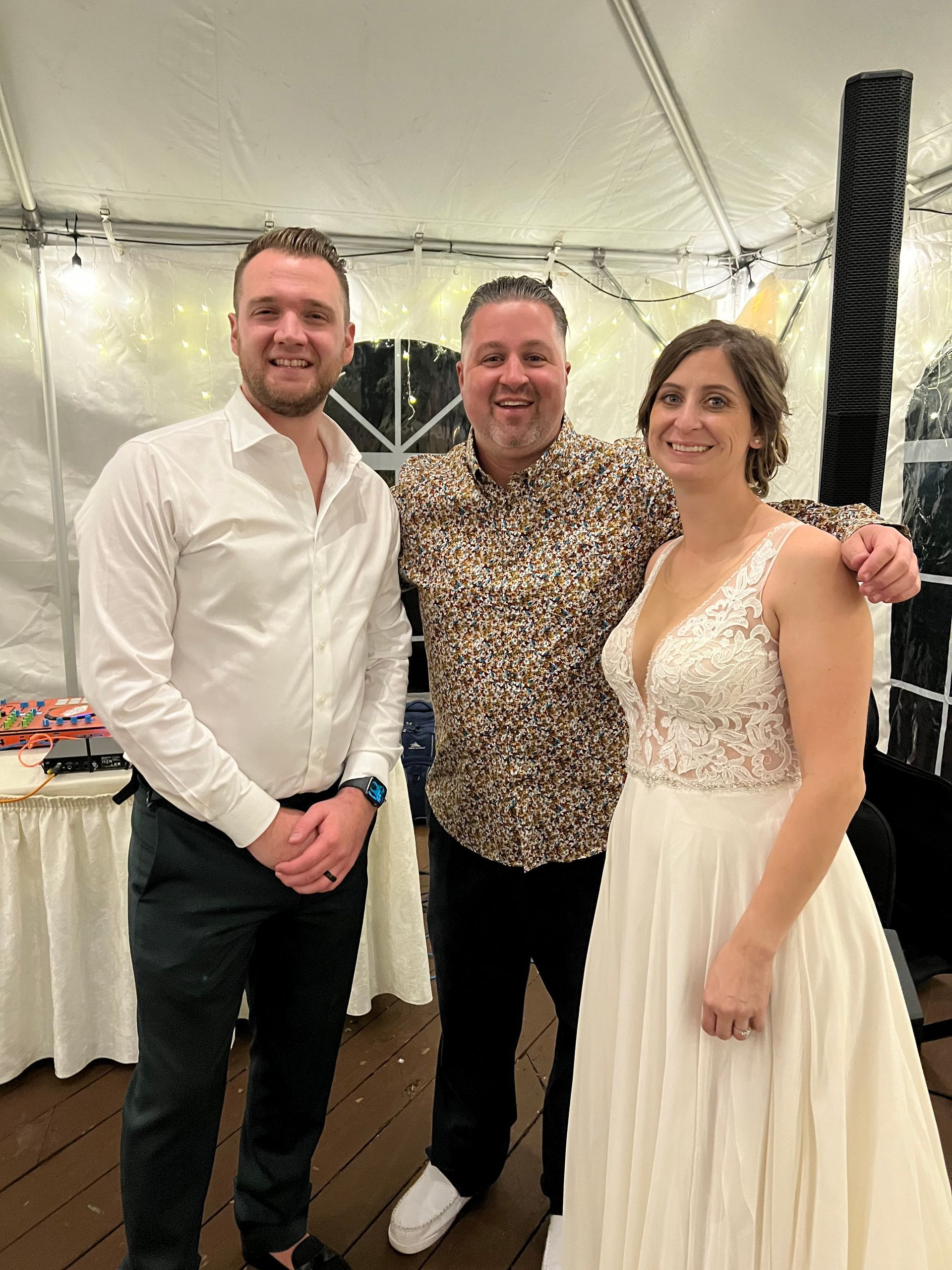 Three people smiling in a tent, the bride in a white dress with the groom in a white button-down shirt next to a man 