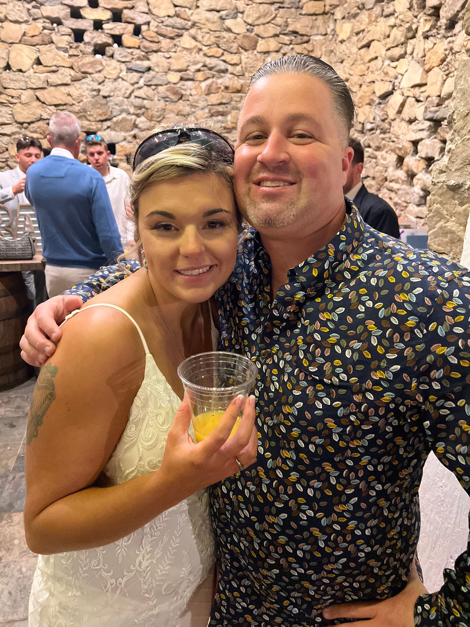 Woman in white dress and man in patterned shirt smile, hugging. Interior with stone walls.