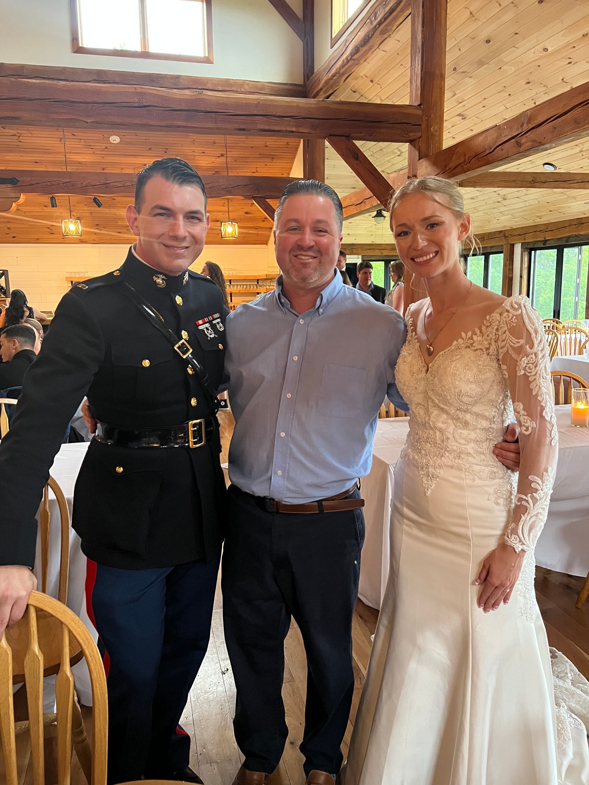 Man in Marine uniform, man in blue shirt, and bride pose together at wedding reception.