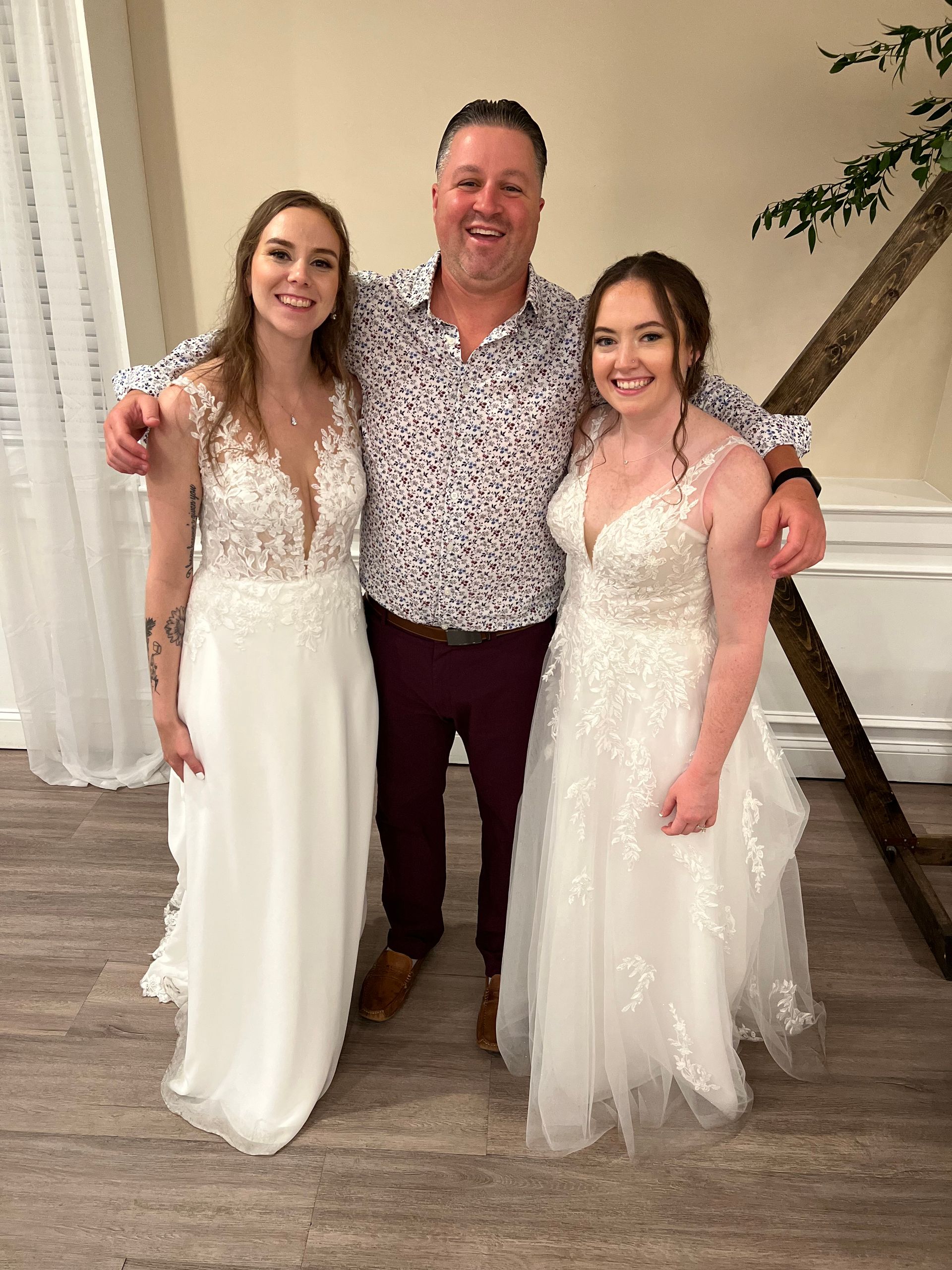 Man smiles, arms around two women in white wedding dresses. Indoors, next to a decorative arch.
