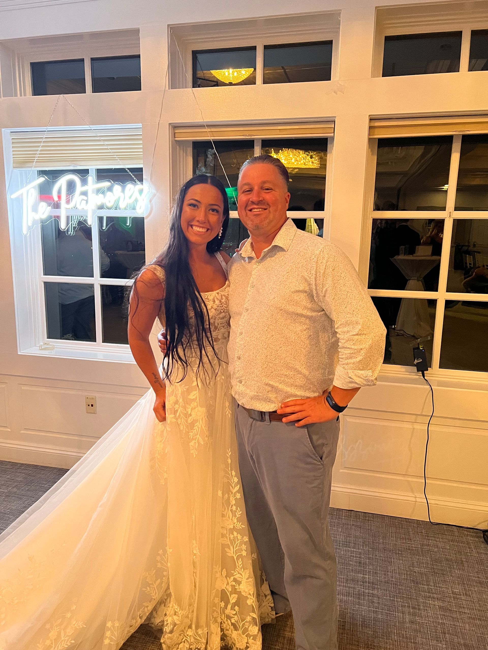 Bride and man pose indoors. Bride in white dress, man in patterned shirt. Windows in background.