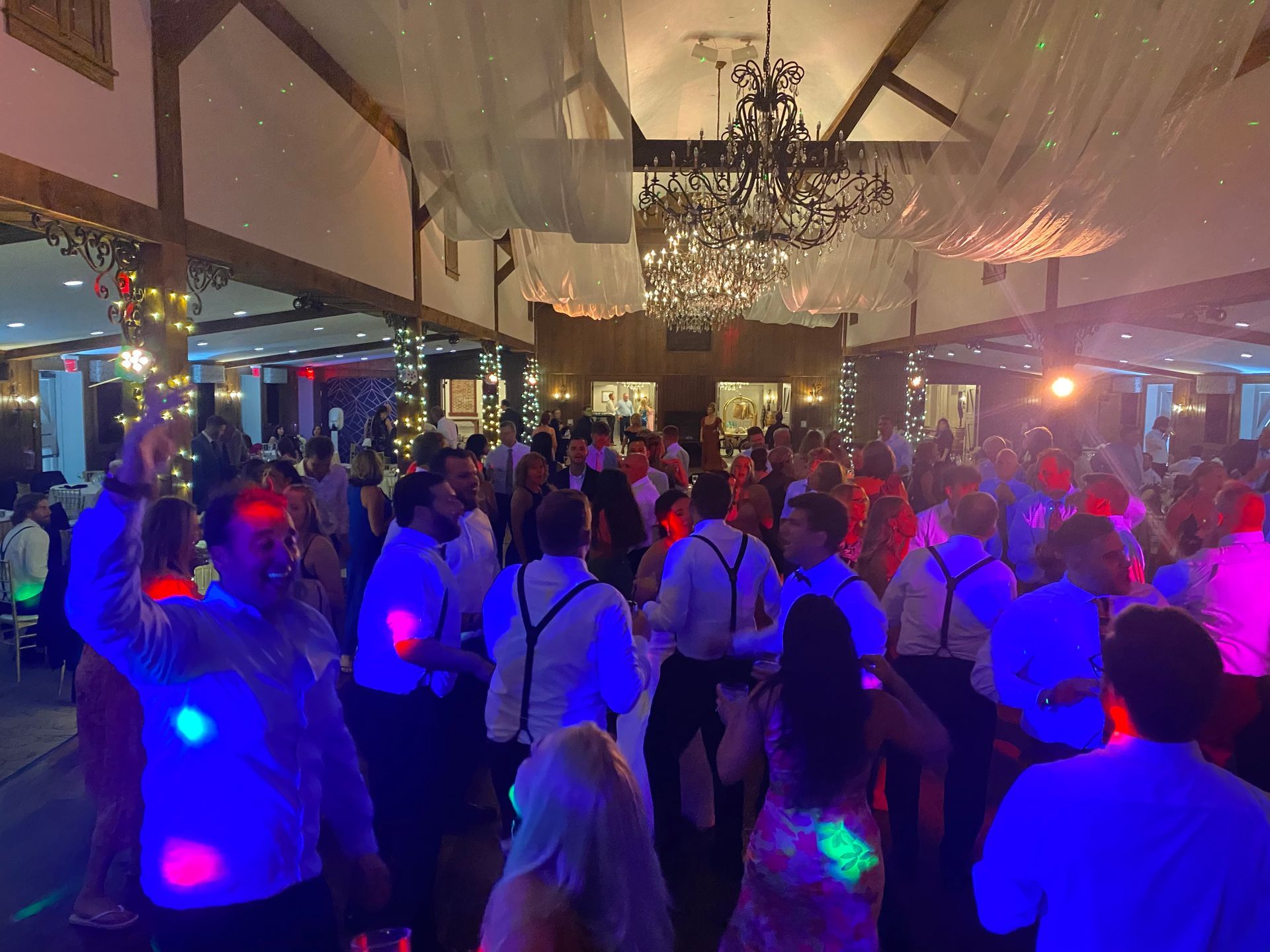 People dancing at a party in a large room with string lights and a chandelier.
