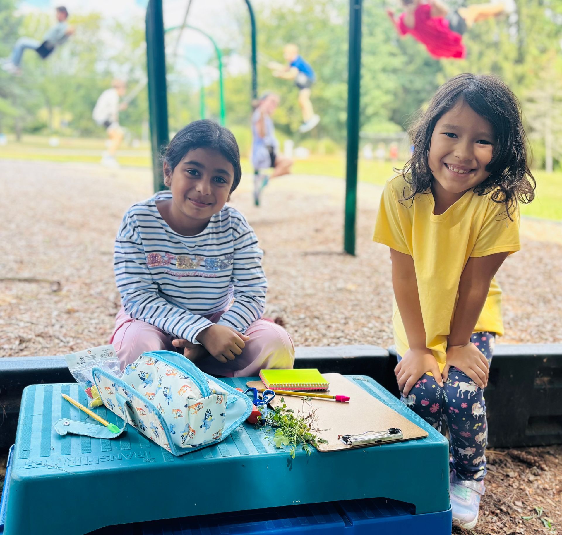 Two children smiling on a playground bench with a toy box and scattered art supplies