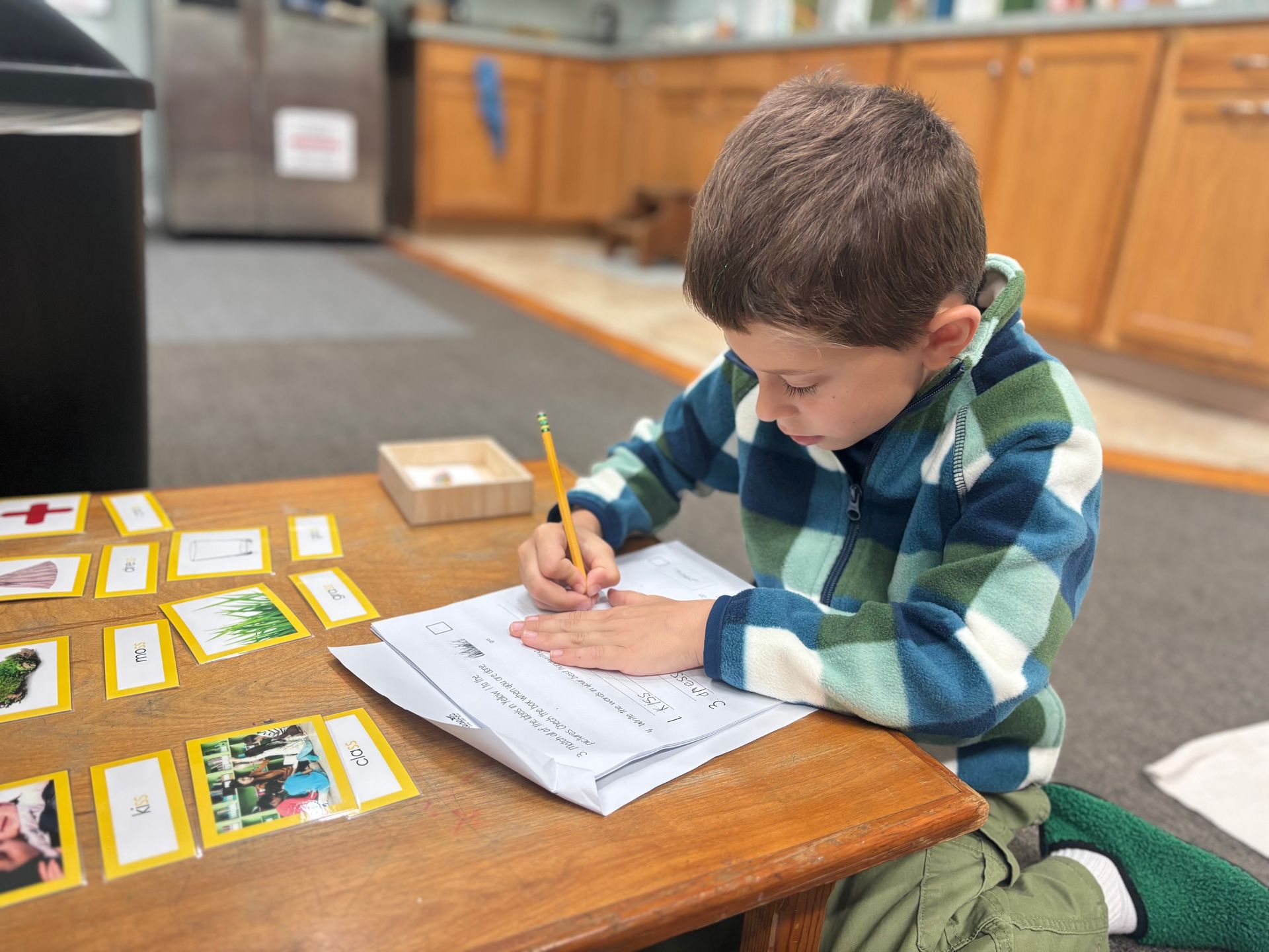 Child in a green plaid shirt writes on a paper at a wooden table, with cards spread beside him.