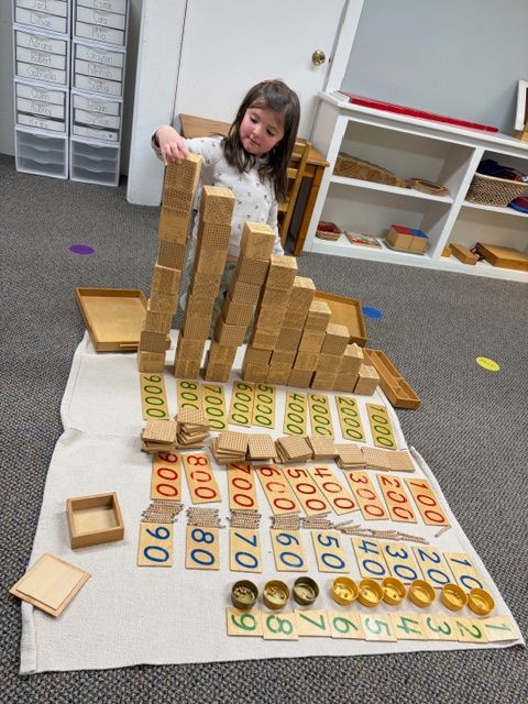 Child building a tall block tower on a classroom floor with large number tiles spread out nearby