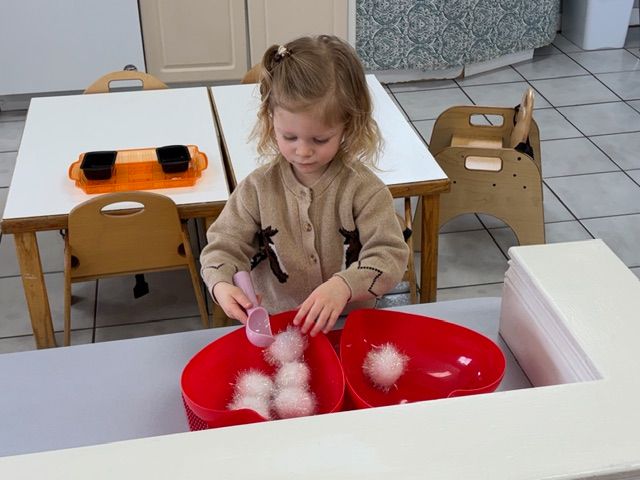 Child playing with cotton balls at a table in a classroom, dropping them into red bowls.