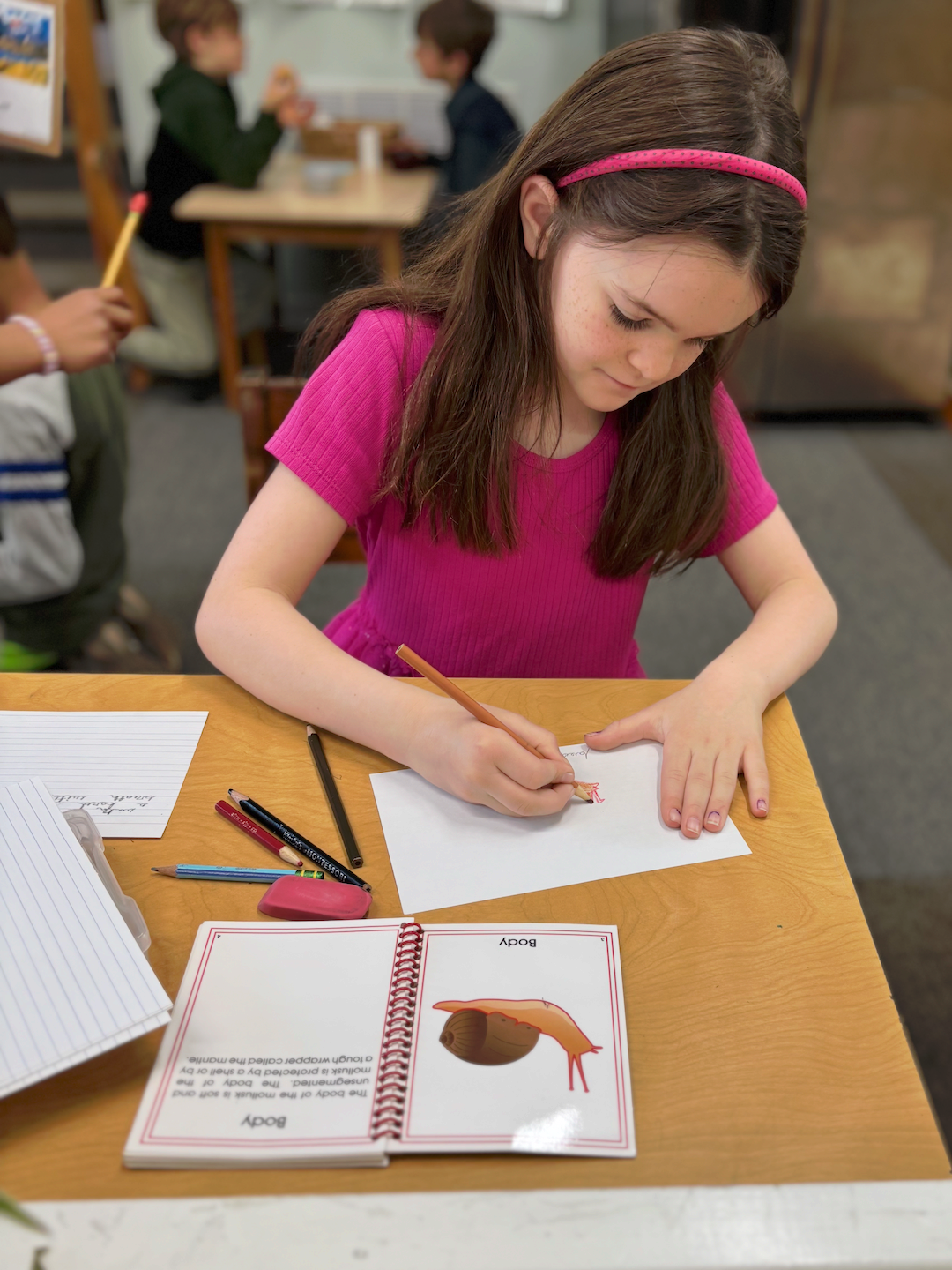 Student in pink shirt writing at a desk in a classroom with worksheets and pencils.