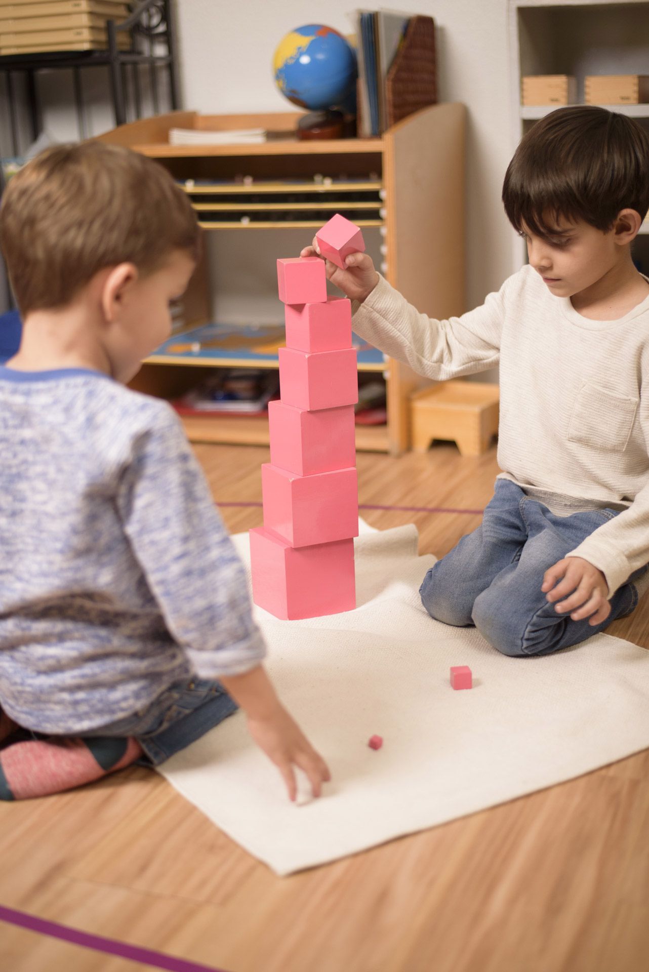 Two Montessori primary students working together on a floor mat to build the Pink Tower sensorial material.