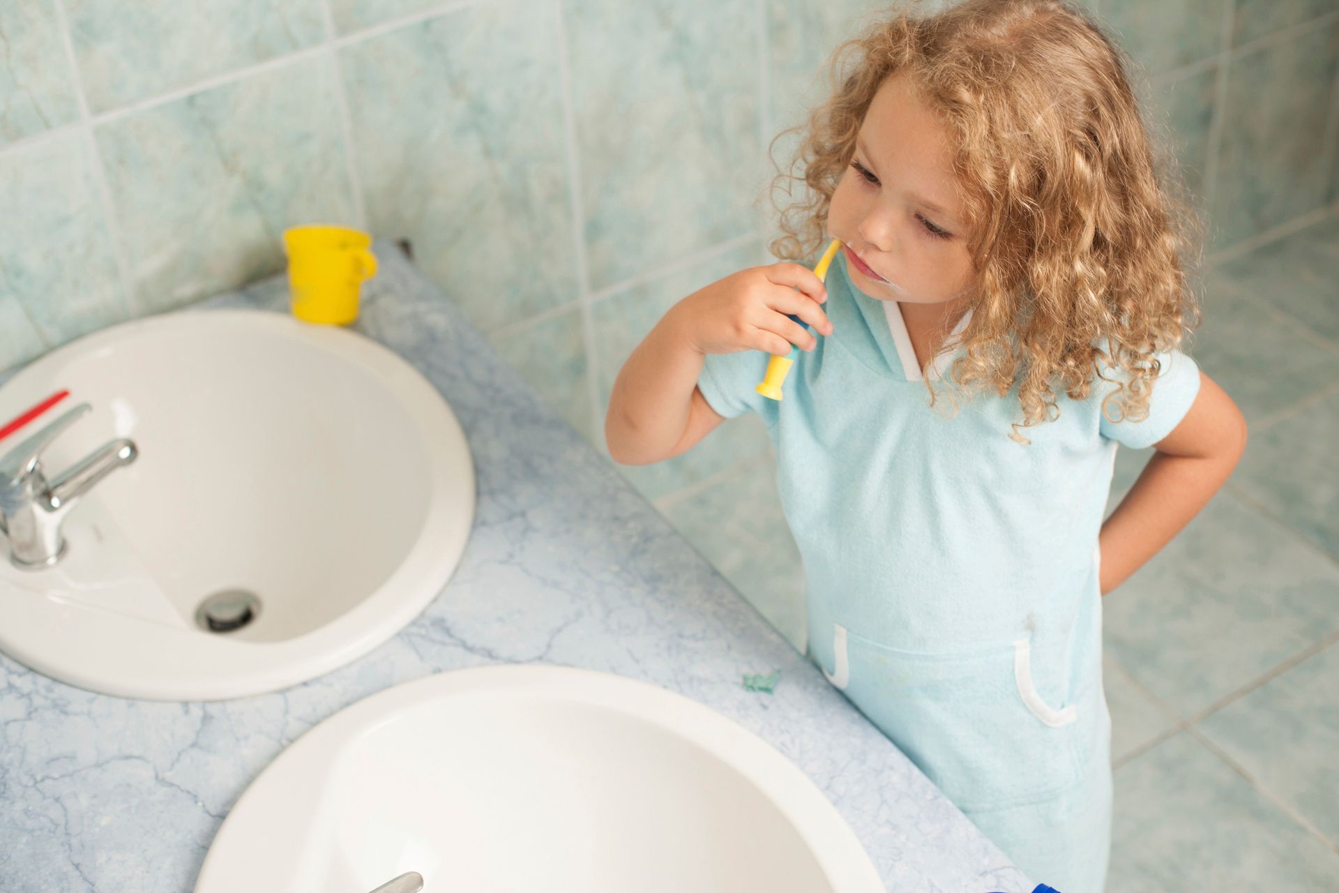 Child in a light blue shirt looking at a toothbrush beside a bathroom sink.