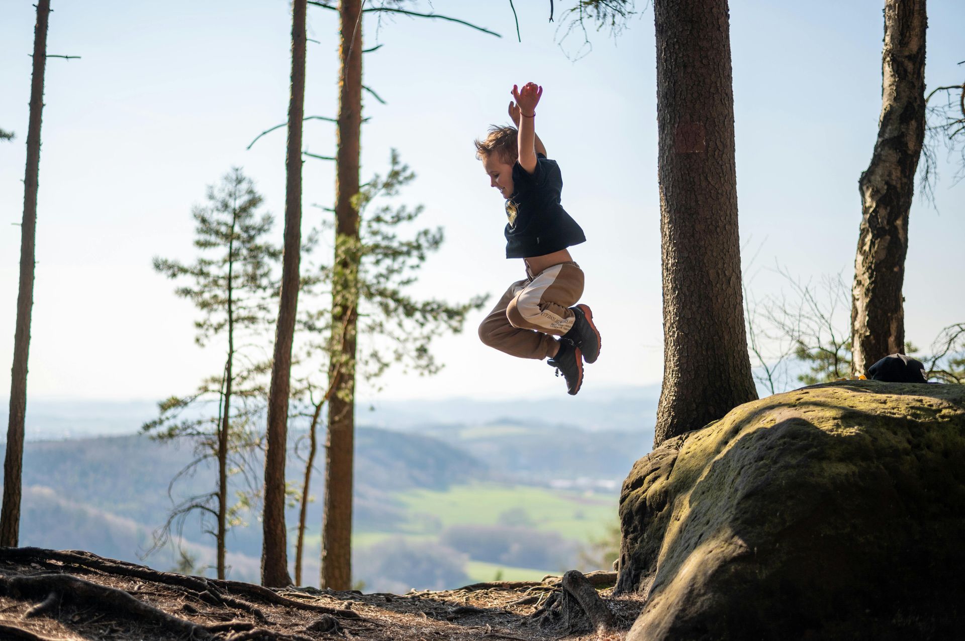 Person jumping between trees on a rocky forest overlook with mountains in the background