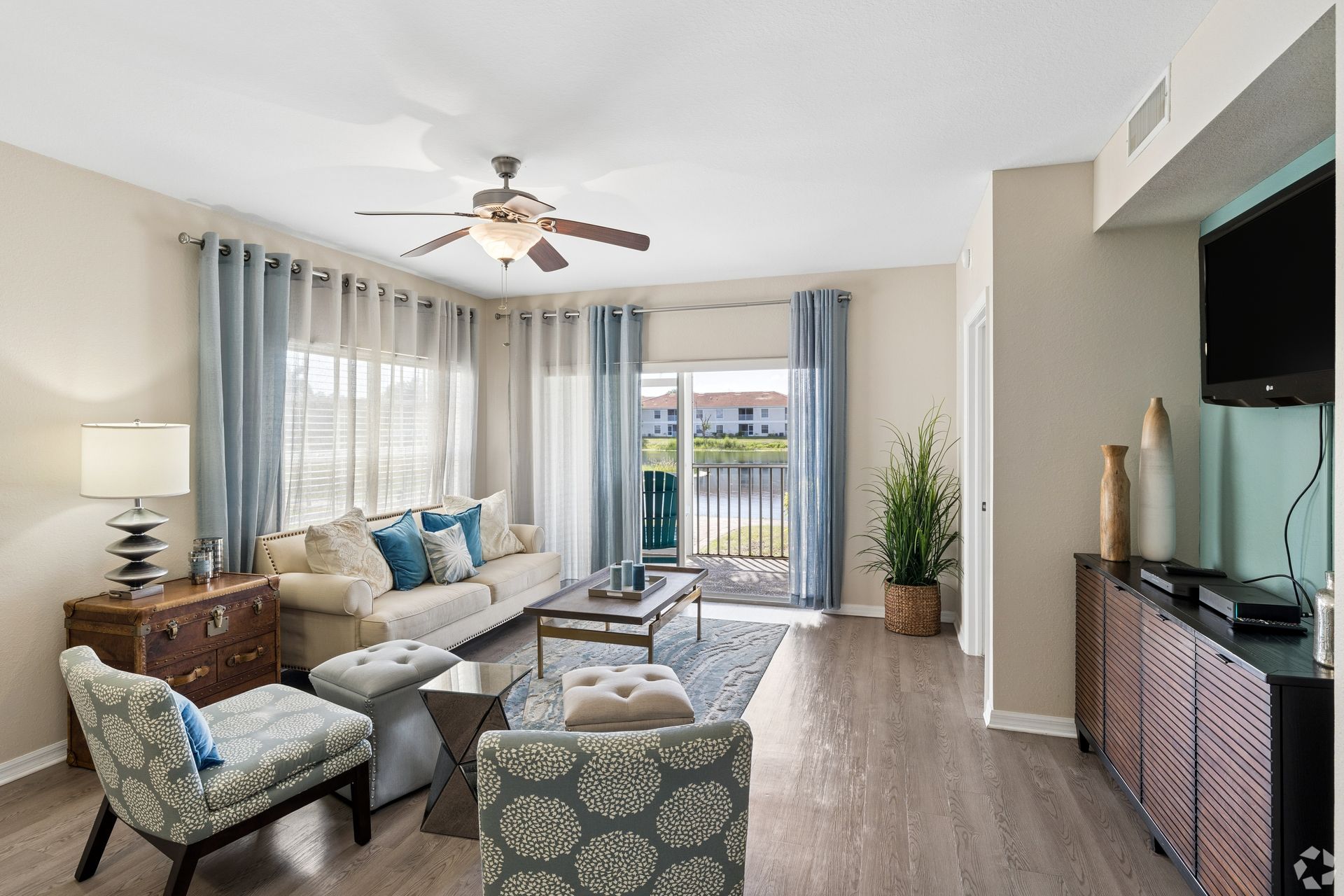 Bright living room with gray sofa, patterned chairs, TV, and balcony doors with sheer curtains