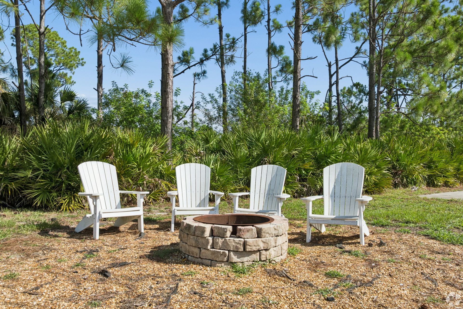 Four white chairs around a stone fire pit in a sunny wooded clearing