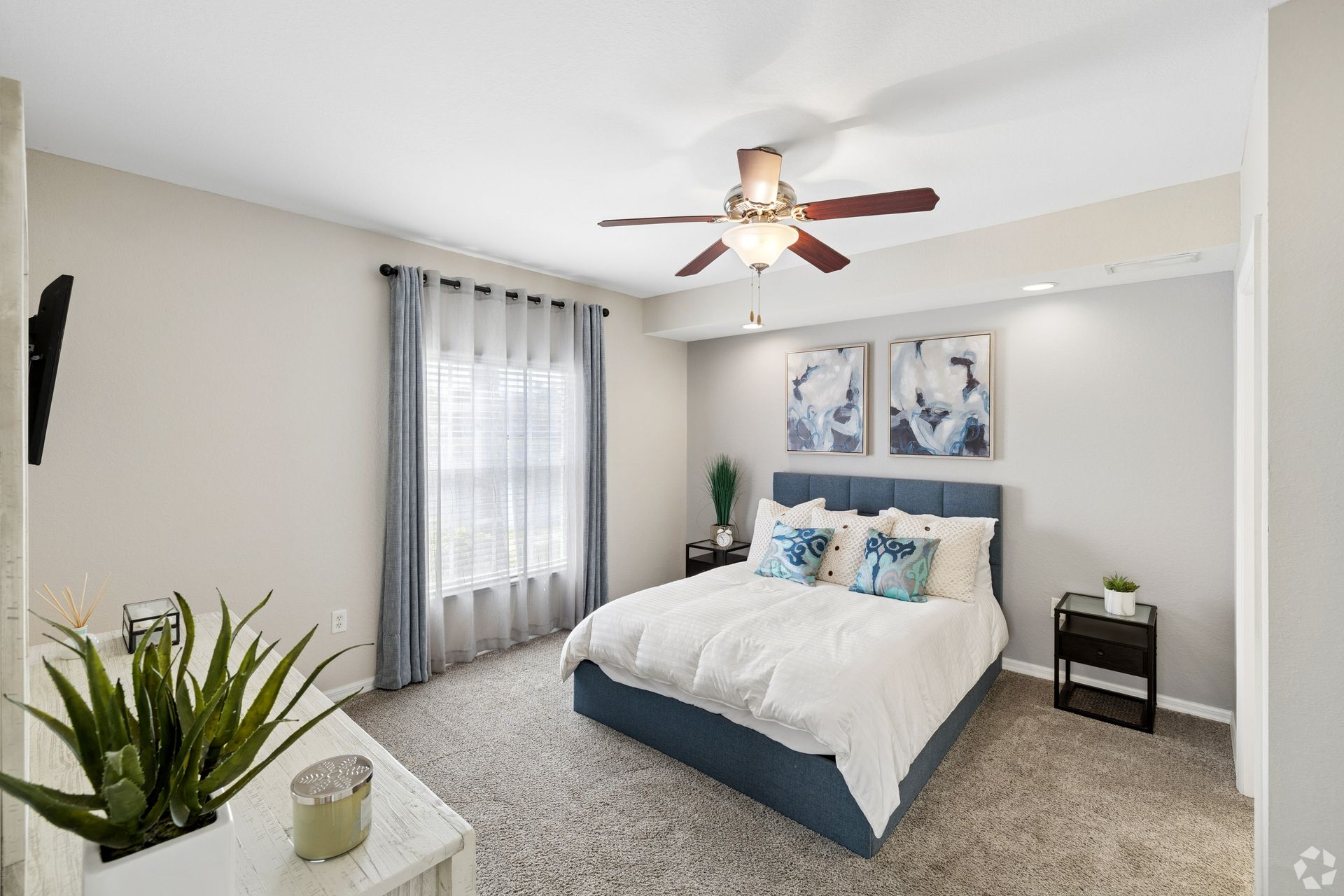 Bright bedroom with a white bed, blue accents, ceiling fan, gray curtains, and framed wall art.