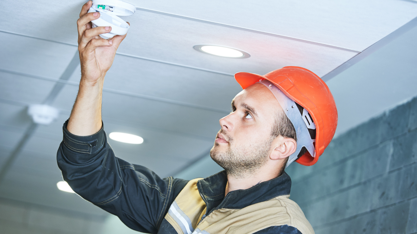 Person in orange hard hat installing a smoke detector on a white ceiling.