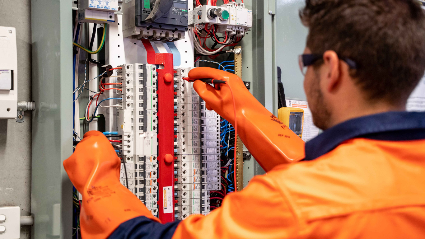 Electrician in orange gloves working on an electrical panel.