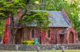 Red stone cottage with ivy and trees, seen behind a low stone wall in a lush green setting