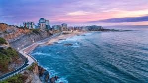 Coastal city skyline beside a curving rocky beach and blue ocean at sunset